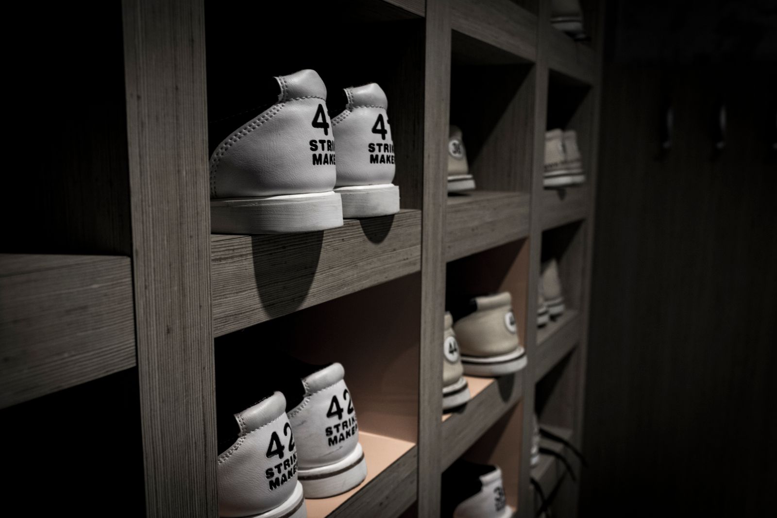Shoes racked in size order on plywood shelving in a dim warehouse unit, one spotlight per bay.